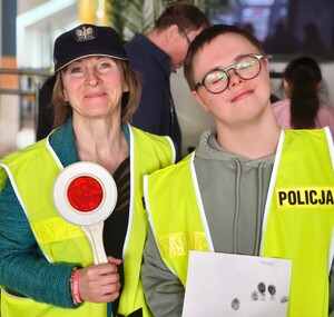 Opiekunka trzymająca tarcze do zatrzymywania pojazdów wraz z podopiecznym w kamizelkach odblaskowych policyjnych.