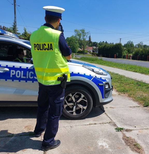 Policjant Wydziału Ruchu Drogowego obok radiowozu dokonuje pomiaru prędkości pojazdu.