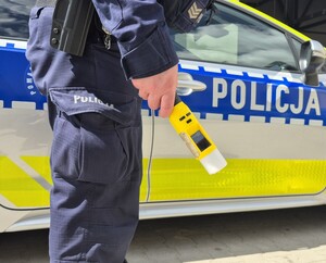 Policjant stojący przy radiowozie trzymający w ręce urządzenie do badania stanu trzeźwości.