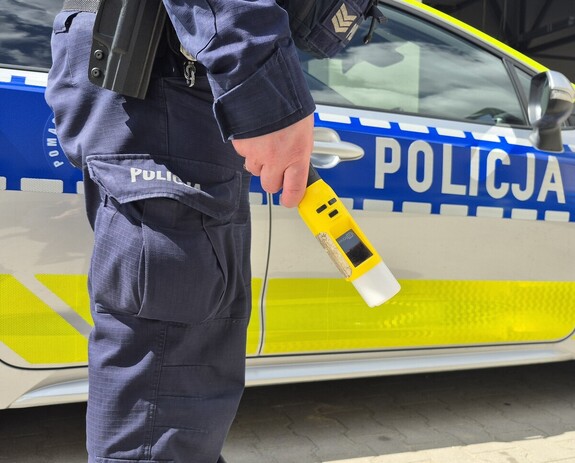 Policjant stojący przy radiowozie trzymający w ręce urządzenie do badania stanu trzeźwości.