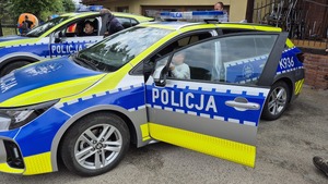 Uczestnik pikniku siedzący za kierownicą radiowozu policyjnego. W tle drugi radiowóz za kierownicą siedzący policjant pokazujący wyposażenie pojazdu.